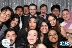 Insha takes a photo with her group of friends from MGMT where there is a silver sparkly wall and everyone is dressed formally, including Insha.