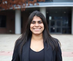 Insha standing on the UBCO campus in front of the science building with orange leaves peeking on the left of the picture. Insha is wearing a black suit.