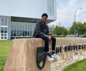 Ifaz is sitting on the University of British Columbia sign in front of the UBCO Commons building wearing all black