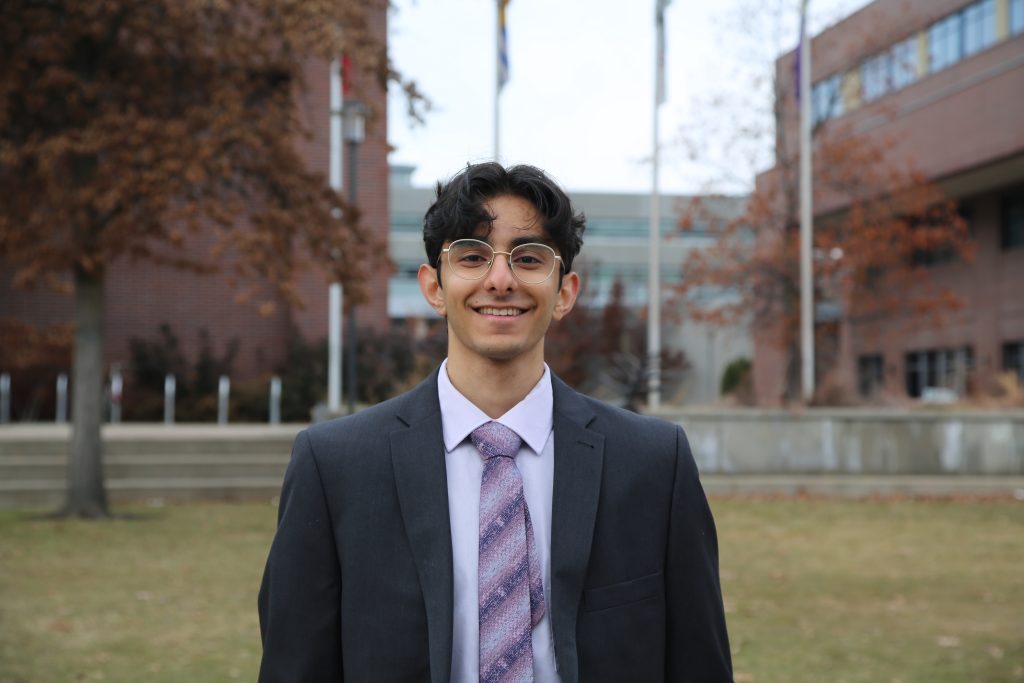 Haider poses for a headshot at the UBCO campus in front of the flags and the Arts building. Haider is dressed formally with a tie.