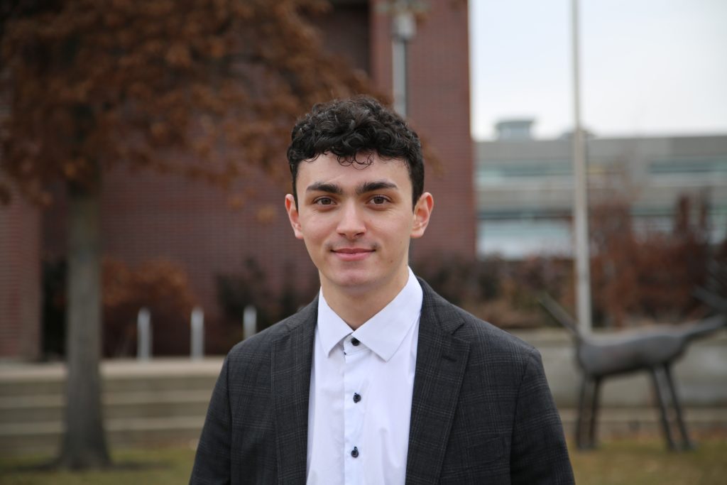 Gianni takes a headshot photo in his professional clothes in the UBCO courtyard with a tree and the art building in the background.