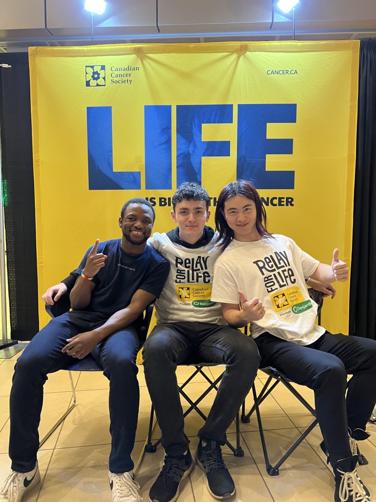 Gianni and his two peers sit on aindividual chair and come together to pose for the photo. The background is a poster of Canadian Cancer Society's poster.