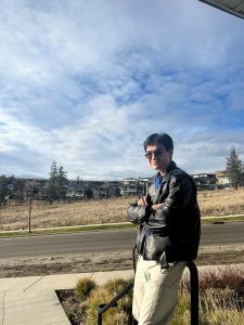 Bao takes a individual photo with a road, dry grass and houses in the background on a nice sunny day with minimal clouds. Bao is in a leather jacket with white pants and shades.