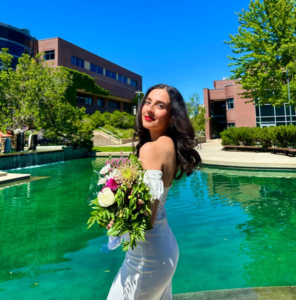 Raneem is wearing a white dress and holding a bouquet of flowers in front of the UBCO courtyard which has the pond filled and it is very sunny and the sky is blue