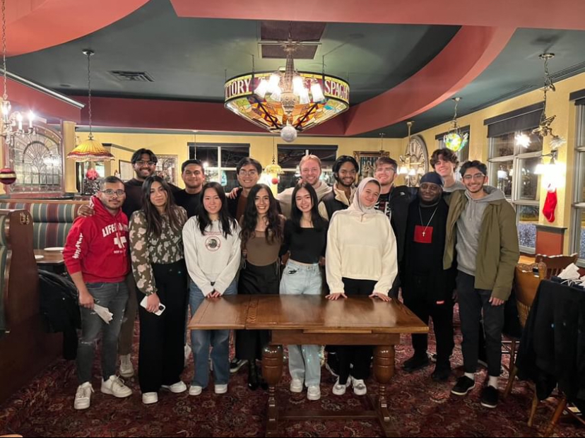 Raneem takes a photo with her MGMT friends at Old Spaghetti factory. There is a wooden table infron of them and they are all standing with a chandelier on top of them.