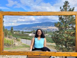 Aashi is wearing a bright blue shirt and standing behind a wooden frame. The background is a wine field with a mountain and lake in the distance. 