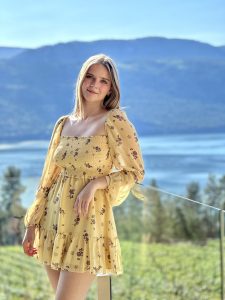 Adriana Velibekova takes a portrait photo weraing a yellow sundress and leans on a glass fence. It is a sunny day and the background is a lake with trees around her in the distance.