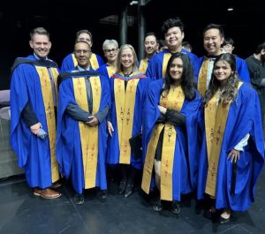 Isabella Bravo takes a photo with the members of the UBC Board of Governors in a deeply lit place with a black background. They are all wearing a black and blue robe.