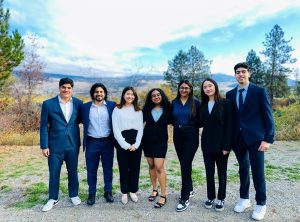 Aashi is dressed up in a blazer, skirt and blue shirt and takes a picture with her friends who are also dressed up. The background is the back of the EME building with a view of the top of the Okanagan valley and blue skies.