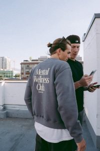 Mahlon and Austin are standing at the top of a building and they are both wearing casual clothes and looking at a phone together.