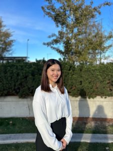 Teresa Mak is wearing a white dress shrit and she is standing in front of a bush and concrete step. There is a tree in the background and blue skies. 