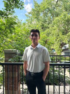 Jacob Baycroft is wearing a polo shirt with blue dress pants and he stands in frotn of a railing and there are lots of trees in the background.