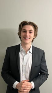 Jarrod Murphy is wearing a black suit with a white dress shirt and takes a photo in front of a grey background