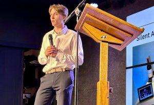 At Jarrod Murphy's Local Business Awareness and Charity event, Jarrod speaks to the crowd wearing a white dress shirt with black dress pants. He is holding a mic and standing beside a podium with a slide show presentation behind him.