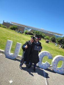 Bravim Paul Saini is dressed up in his graduation gown and hat and takes a picture in front of a white UBCO sign with his graduation degree. He takes a picture with his friend on a very sunny day and the photo is a bit slanted.