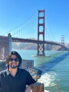 On a sunny day with no clouds, Bravim stands at the edge of a railing where he is in front of the golden gate bridge. There is a wide body or water beside him. Bravim is wearing black and sun glasses.