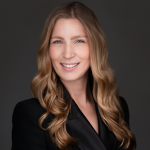 Kelly Davidson takes a close up headshot with a dark grey background. Kelly is wearing a black blazer.