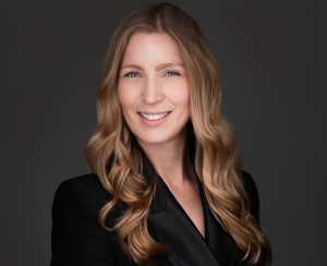 Kelly Davidson takes a close up headshot with a dark grey background. Kelly is wearing a black blazer.