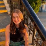 Grace Ingalls takes a golden hour picture wearing a black top and jeans. She is sitting on concrete stairway with black railings.