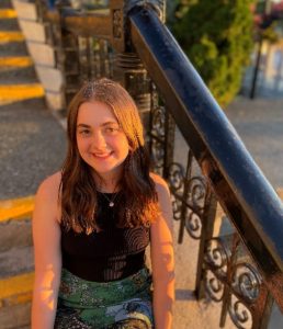 Grace Ingalls takes a golden hour picture wearing a black top and jeans. She is sitting on concrete stairway with black railings.