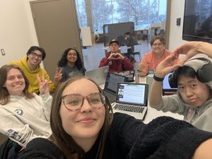 Lamay Boshoff takes a selfie with her friends in a Commons study room. There are many people doing cute hand poses and computers in the center. 