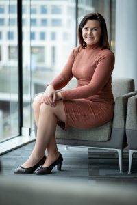 Catherine Melville is sat on a chair, dressed in a dark orange dress with black heels. Beside her is a big window that has a view of another building in the distance.