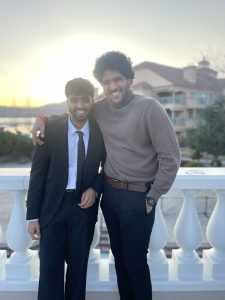 Akshat Porwal is all dressed up and takes a picture with his friend. They are standing in front of a white railing and the background includes an apartment and the bright sun.