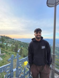 Akshat Porwal takes a picture at the top of a mountain in front of a railing. Behind him are moutains, trees and a beautiful sunset with a bit of orange. 